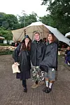 Tre festivalgæster i sorte regnslag står foran telt og borde på udendørs festivalplads.