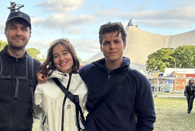 Tre personer står sammen på en festivalplads omgivet af telte og græs