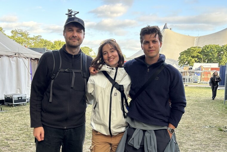 Tre personer står sammen på en festivalplads omgivet af telte og græs