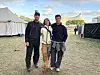 Tre personer står sammen på en festivalplads omgivet af telte og græs