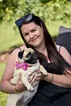 Maja fra tv-programmet holder en mops med pink sløjfe i skødet i en grøn have.
