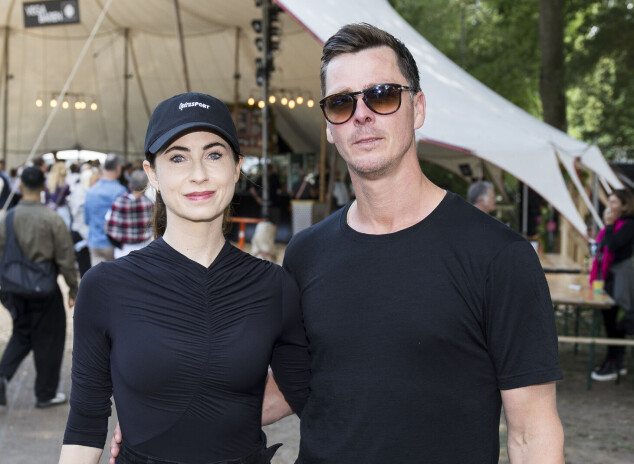 Par står arm i arm foran telt og langborde på udendørs festivalområde.