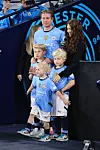 Kevin De Bruyne står på banen med sin familie i Manchester City-trøjer foran stor klublogo-baggrund.