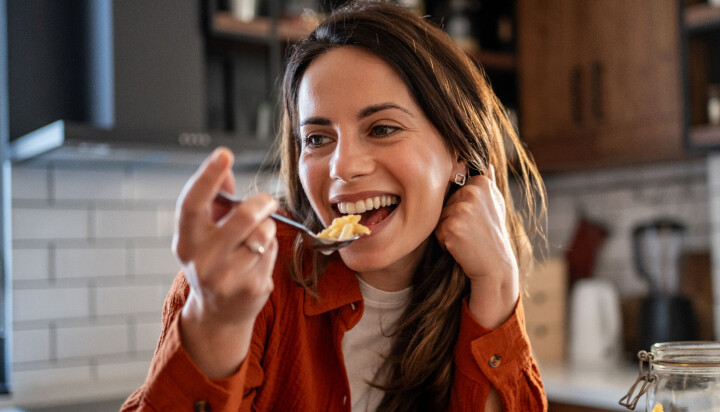 Smilende kvinde spiser morgenmad ved et køkkenbord med glasbeholdere med müsli.