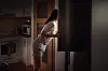 Kvinde i pyjamas læner sig ind i et åbent køleskab i et mørkt køkken om natten