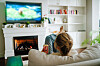Kvinde med håret i knold ser tv på sofa foran tændt pejs i lys stue.