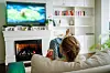 Kvinde med håret i knold ser tv på sofa foran tændt pejs i lys stue.