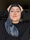 Person med silkehat og sort bluse poserer med hænderne på hovedet indendørs