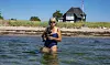 Pia Kjærsgaard i badedragt står i havet ved Enø med en hund foran et sommerhus.