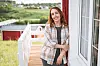 Smilende kvinde står op ad et rødt hus på en træterrasse med udsigt over grønt landskab.