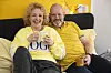 Mette Vibeke og Michael Ramgil Stæhr sidder i seng med krus foran en gul væg med lystkæde.