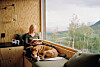 Kvinde læser ved stort vindue i træhytte med hund liggende ved siden af.