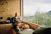Kvinde læser ved stort vindue i træhytte med hund liggende ved siden af.
