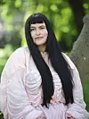 Emilie Foli med langt mørkt hår i lyserød kjole sidder ved et træ i en grøn park.