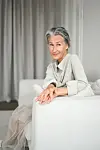 Ældre kvinde med gråt hår sidder afslappet på lys sofa i rolig stue.