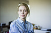Kvindeportræt i hjemlig stue, hvor en midaldrende kvinde i denimskjorte ser alvorligt mod kameraet