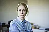 Kvindeportræt i hjemlig stue, hvor en midaldrende kvinde i denimskjorte ser alvorligt mod kameraet