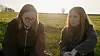 To unge kvinder sidder i græsset på en solbelyst mark og ser eftertænksomme ud