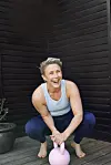 Smilende kvinde i træningstøj sidder på terrasse og holder en lyserød kettlebell.