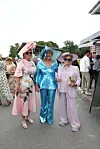 Tre kvinder i farverige festoutfits og store hatte står sammen ved et udendørs arrangement.