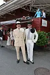 To elegant klædte mænd står foran en rød træbygning ved et udendørs event.