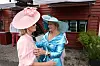To ældre kvinder i farverige kjoler og hatte hilser på hinanden foran en rød træbygning.