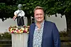 Mand i jakkesæt ved skulptur med guitar på piedestal pyntet med roser under grønne træer.