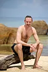 Mand i badebukser sidder på en træstamme på sandstrand ved havet