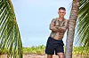 Tatoveret mand står med korslagte arme ved en palme på en strand