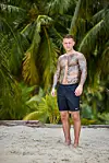 Tatoveret mand i sorte Nike-shorts står på sandstrand foran grønne palmer