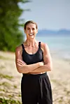 Smilende kvinde i sort sportstøj står med korslagte arme på en strand.