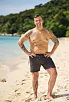 Mand i badeshorts står på solrig sandstrand foran turkis hav og grøn kystlinje.