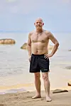 Mand i sorte badebukser står på sandstrand foran stille hav
