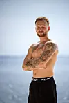 Tatoveret mand står ved havet med korslagte arme i solen.