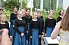 Børnekor i blå nederdele og sorte bluser synger udendørs foran en dirigent
