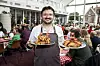 Umut Sakarya står på Flæskekroen med to fade stegt flæsk foran en fyldt spisesal.