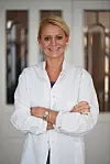 Kvinde i hvid kittel står med korslagte arme i en lys indendørs gang.