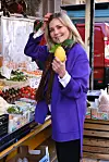 Kvinde i lilla frakke holder en stor citron ved en grøntsagsbod på markedet