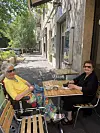 To ældre kvinder sidder og taler ved et bord på en solrig fortovscafé.