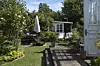 Solrig kolonihave med træterrasse, havemøbler, blomsterbede og lille hvidt havehus i baggrunden.