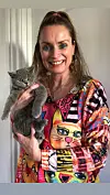 Kvinde i farverig bluse holder en grå British Shorthair-killing foran en hvid dør.