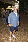 Ældre mand i denimskjorte og shorts går på festivalplads med mennesker og lys i baggrunden.