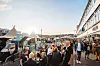 Mennesker sidder ved borde og spiser ved en travl havnepromenade i aftensol.