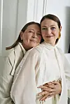 Ældre og yngre kvinde står tæt og omfavner hinanden i en lys stue.