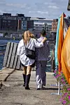 To personer fra Temptation Island går arm i arm langs en havnepromenade med bygninger i baggrunden.