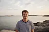 Smilende mand står på klipper foran roligt hav i aftensol.