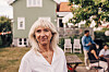 Ældre kvinde i hvid skjorte står i en have foran et lyst træhus med gæster i baggrunden.