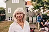 Ældre kvinde i hvid skjorte står i en have foran et lyst træhus med gæster i baggrunden.