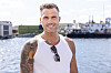 Smilende mand fra Temptation Island står ved en havn med vand og skibe bag sig.
