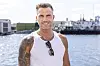 Smilende mand fra Temptation Island står ved en havn med vand og skibe bag sig.